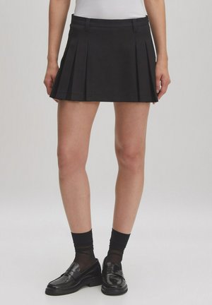 Black pleated mini skirt with a smooth texture, featuring a waistband and side pockets. Worn with dark socks and loafers.