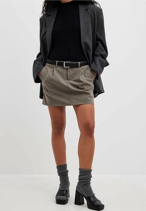 Grå blazer, sort ribbet top, khaki plisseret miniskirt, sort bælte, grå strikstrømper, sorte plateauhæle. Ensfarvet baggrund.