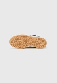 The underside of a sneaker shows a textured rubber sole with a zigzag pattern, featuring a beige color and a black shoe upper partially visible.