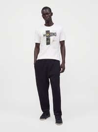 Homme debout les mains dans les poches, portant un t-shirt blanc Moschino graphique, un pantalon noir et des baskets noires sur un fond clair uni.