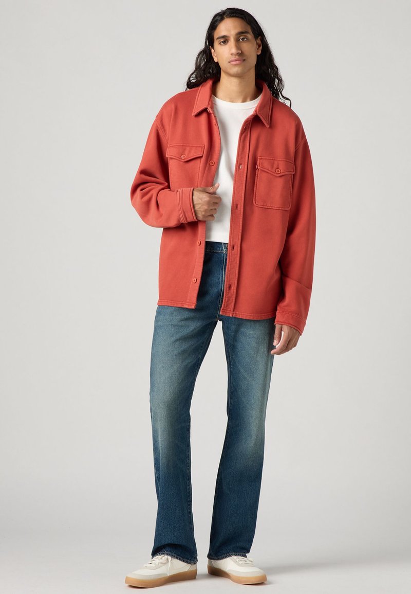 Röd bomullsblandad skjortjacka med två bröstfickor, bärs �över en vit t-shirt, kombinerad med blå jeans och krämfärgade sneakers.