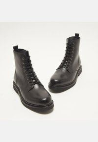 Čierne kožené kotníkové boots s hladkým povrchom, okrúhlou špičkou a plochou gumovou podrážkou. Obsahujú šnurovanie a pútko na päte.
