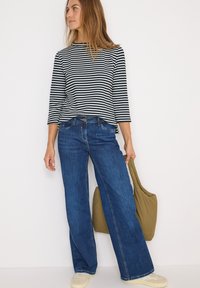 Maglietta a maniche lunghe a righe bianche e nere abbinata a jeans blu a gamba larga. Il modello tiene in mano una borsa a tracolla verde oliva. Sneakers di colore crema presenti.
