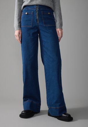 Denim wide-leg pants in deep blue, featuring a gold zipper and button accents, with large back pockets and a straight cut at the hem.