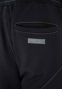 Čierne teplákové nohavice s textúrovaným povrchom, s zadnou kapsou a kontrastným svetlomodrým prešívaním. Štítok znie "MULTIPLY APPAREL."