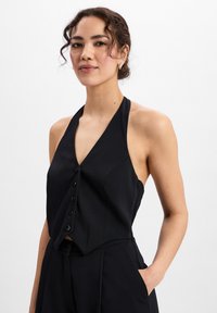 Schwarzes Haltertop mit V-Ausschnitt, ausgestattet mit einer Knopfverschluss-Front und einem strukturierten Design, kombiniert mit einem eleganten schwarzen Outfit.