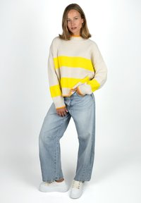 Beiger Pullover mit zwei leuchtend gelben horizontalen Streifen, mit gerippten Bündchen und Kragen. Kombiniert mit hellblauen weit geschnittenen Jeans und weißen Turnschuhen.