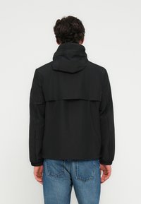 Μαύρο αδιάβροχο μπουφάν με κουκούλα, σχεδιασμένο με άνετη εφαρμογή, ελαστικά μανίκια και λεπτομέρεια πίσω στον ώμο. Φορεμένο πάνω από ανοιχτόχρωμα τζιν.