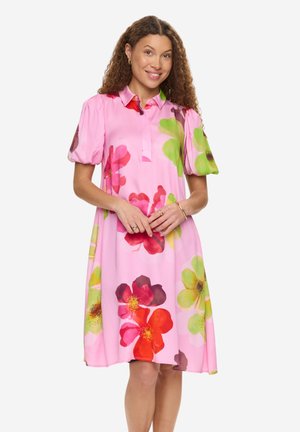 Kvinde med krøllet hår iført en lyserød kjole med store farverige blomsterprint, der smiler med hænderne blidt foldet foran sig.