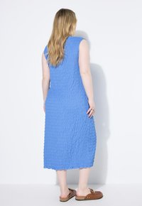 Vestido azul hasta la rodilla, hecho de una tela texturizada con un dobladillo volante, con un diseño sin mangas. Combinado con zapatos marrones tipo mocasín.