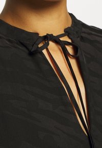 Top negro con un patrón texturizado, con un escote tipo ojal asegurado por lazos negros y un cuello redondeado. Acabado de tela suave.