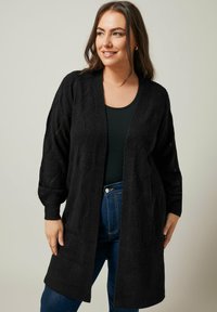 Schwarzer langer Cardigan mit einem strukturierten Strickmuster, offener Front und Dreiviertelärmeln, kombiniert mit einem schwarzen Oberteil und blauen Jeans.