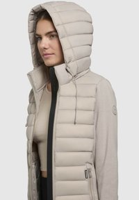 Beige quiltet pufferjakke med hætte, med lynlås foran, sidelommer og struktureret ærme materiale for ekstra varme.
