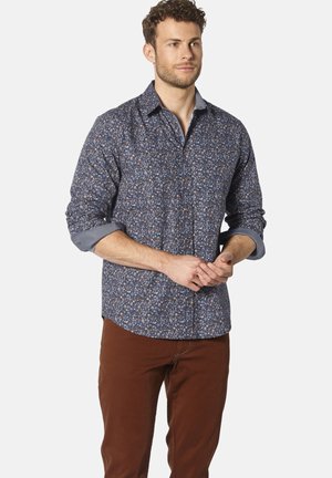 Blaues Blumenhemd mit Button-Down-Kragen, langen Ärmeln und hochgekrempelten Manschetten; es zeigt kleine Blumen auf einem dunklen Hintergrund. Getragen mit braunen Hosen.