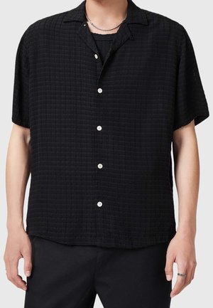 Man wearing black textured short-sleeve button-up shirt over a black t-shirt, with silver chain necklace and black pants.