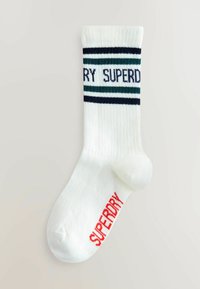 Hvide ribbede sokker med "SUPERDRY" i rødt på foden og sorte og grønne striber med delvist "SUPERDRY"-tekst nær toppen af ribbenet.
