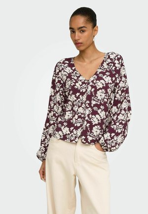 Vrouw die een paarse blouse met lange mouwen en witte bloemenprint draagt en crème hoogtaille broek, staat met één hand in de zak.