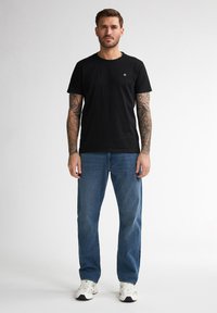 Zwarte katoenen T-shirt met een ronde halslijn en een klein logo op de borst, gecombineerd met blauwe spijkerbroek en witte sportschoenen.