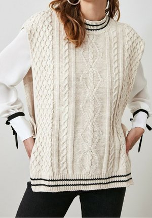 Kvinde iført en cremefarvet, kabelstrikket ærmeløs sweatervest med sortstribet krave og kant over en hvid bluse med sorte båndmanchetter.