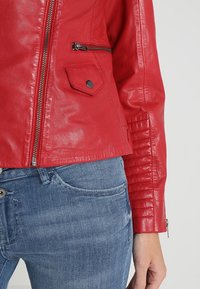 Giacca in pelle rossa con zip frontale, tasche laterali e texture a coste sulle maniche; indossata sopra jeans in denim azzurro chiaro.