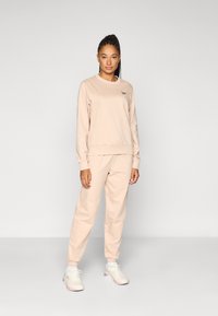 Gaiši bēšs džemperis un atbilstoši sporta bikses, mīksts audums, apaļš kakls, ribots aproces un vidukļa josla, ar smalku logotipa detaļu krūtīs.