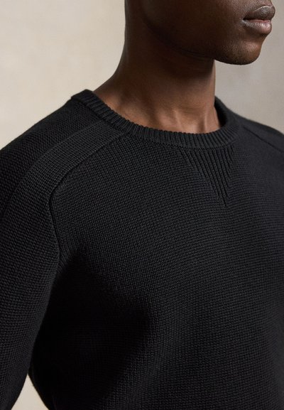 Gros plan d’une personne portant un pull en maille noire texturée avec un col rond et des détails côtelés aux épaules et au cou.