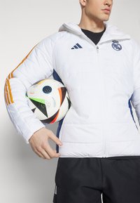 Giacca puffer bianca con accenti blu, caratterizzata da strisce arancioni sulle maniche, chiusura con zip e cappuccio, sostenuta contro un pallone da calcio colorato.