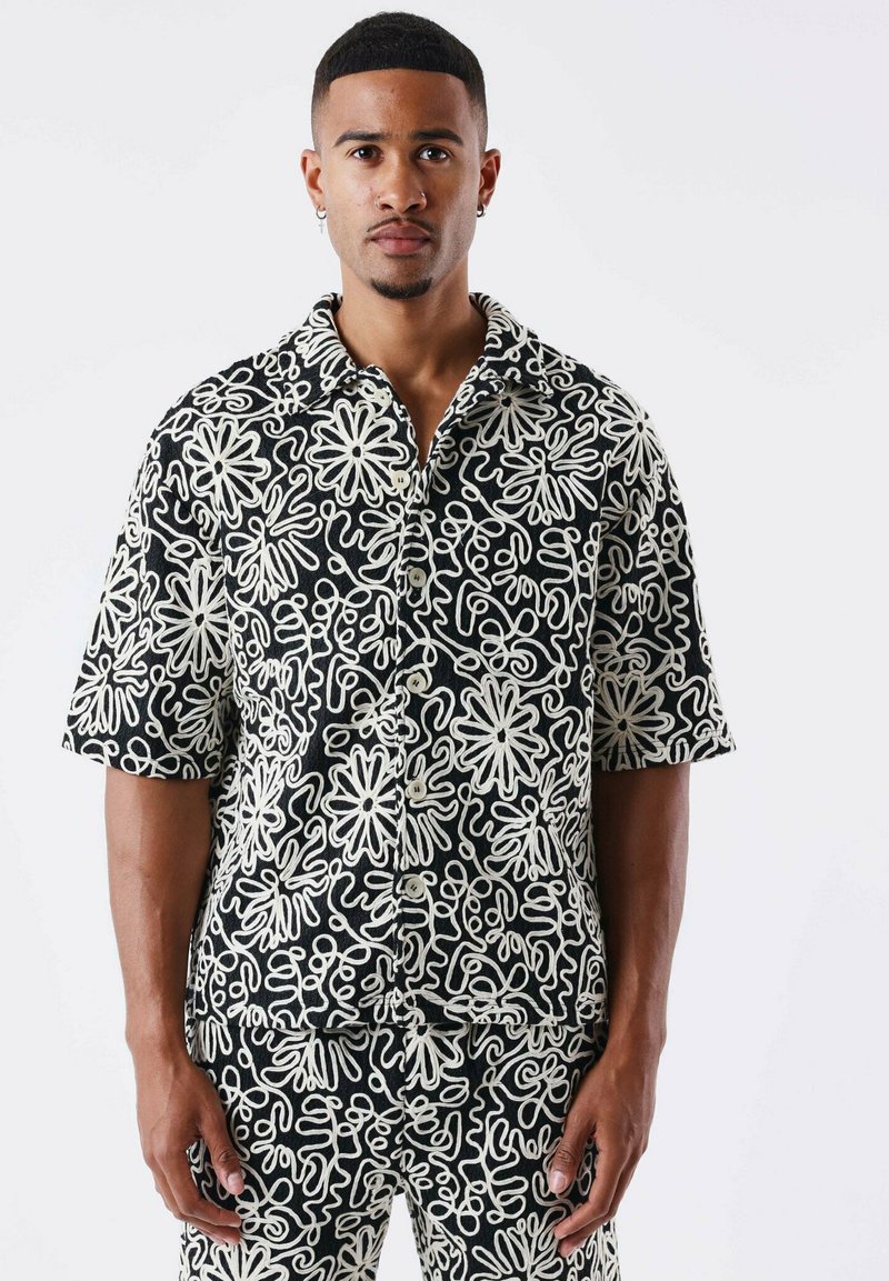 Camisa de manga corta en negro con un patrón floral blanco, con cierre de botones en la parte delantera y un corte relajado. Diseño de tela texturizada distintiva.