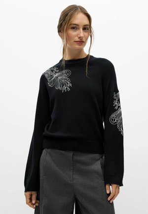 Modèle portant un pull noir orné de motifs floraux blancs sur l’épaule et la manche, associé à un pantalon gris, sur un fond blanc uni.