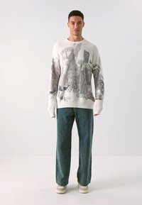 Weißer gerippter Pullover mit einem grauen grafischen Druck, kombiniert mit türkisfarbenen Jeans und beigen Sneakers. Das Outfit hat eine entspannte Passform und einen lässigen Stil.