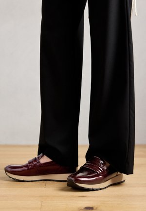 Chaussures à enfiler en cuir verni bordeaux avec une semelle plate et un pantalon noir, présentant des coutures discrètes et une finition lisse.