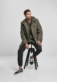 Olivno zelena parka s kapuco obrobljeno z krznom, v kombinaciji z vojaško srajco in črnimi hlačami, nošena z črnimi supergami. Lesena stolica kot podpora.