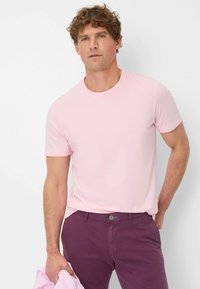 Maglietta rosa chiaro a maniche corte con colletto rotondo, abbinata a pantaloni viola scuro, tenuti con una mano. Tessuto dalla texture liscia.