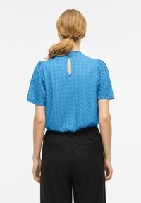 Blouse bleue à manches courtes avec un motif zigzag texturé, encolure ronde et détail fente dans le dos, associée à un pantalon noir.