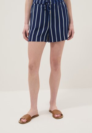 Femme portant un short bleu marine avec des rayures verticales blanches et jaunes, associé à des sandales slides marron sur un fond clair uni.