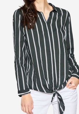 Femme portant un chemisier noir à manches longues avec des rayures blanches verticales et un nœud à l’avant, associé à un pantalon blanc.