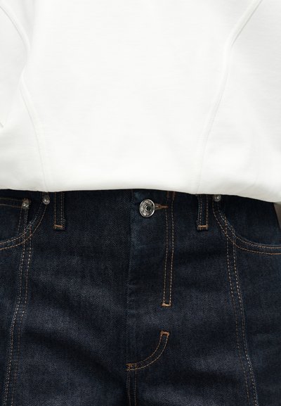 Κοντινό πλάνο σκούρων μπλε τζιν παντελονιών με μεταλλικό κουμπί και πορτοκαλί ραφές σε αντίθεση, κάτω από λευκή μπλούζα με κουκούλα.