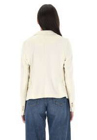 Blazer bianco con colletto, due tasche anteriori e polsi con bottoni, abbinato a jeans dritti blu. Tessuto liscio e vestibilità sartoriale.