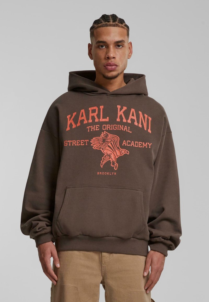 Sudadera marrón con un bolsillo canguro en el frente, con un estampado gráfico naranja que incluye un diseño de águila y texto. Material de algodón, corte relajado.