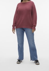 Pull tricoté couleur rose avec un motif texturé, associé à un jean bleu clair à jambes droites avec des fentes latérales et des baskets foncées.