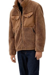 Brun sherpa fleecejakke med lynlås, to brystlommer med klap og to sidelommer med lynlås, båret over mørke jeans.