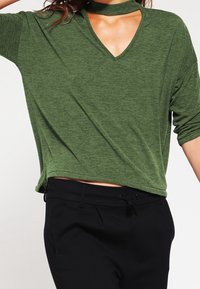 Grünes V-Ausschnitt-Top mit einem Choker-Detail, aus weichem Stoff, mit kurzen Ärmeln und lockerer Passform. Kombiniert mit schwarzen Hosen.
