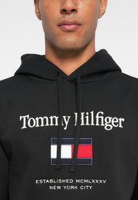 Schwarzer Hoodie mit weiß gesticktem Text "Tommy Hilfiger" und einem rechteckigen Logo mit roten, weißen und blauen Elementen. Gemütliche Stoffstruktur.