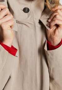 Beżowy płaszcz z kołnierzem, zapinany na guziki, z widocznym przeszyciem. Czerwony detal przy mankietach widoczny z rękawów. Gładka tekstura materiału.
