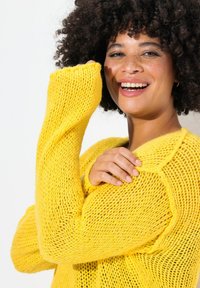 Gelber gestrickter Pullover mit einer lockeren, offenen Maschenstruktur, langärmlig und mit strukturierten Akzenten. Das Modell lächelt und berührt leicht das Kleidungsstück.