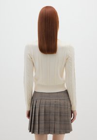Cremfarbener Strickpullover mit vertikalen Rippungen und Kabelakzenten, kombiniert mit einem karierter Faltenrock in Braun- und Orangetönen.