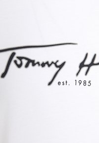 T-shirt in cotone bianco con un logo nero di firma che recita "Tommy" e "est. 1985" in un carattere contrastante. Texture liscia.