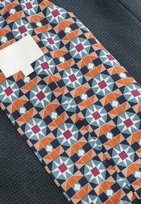 Tekstil med et geometrisk mønster i orange, blå og creme med en navy blå tekstureret accent. Inkluderer et hvidt etiketafsnit.