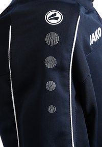 Veste de sport bleu marine avec accents blancs, ornée d'un logo circulaire et de motifs décoratifs en pointillés le long de la couture de l'épaule.