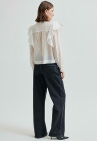 Kobieta ubrana w przezroczystą białą bluzkę z falbankami na ramionach, czarne spodnie o szerokich nogawkach i czarne buty na obcasie w szpic, stojąca tyłem na jednolitym tle.